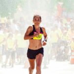 A female athlete running in a marathon with a crowd cheering on a sunny day.