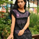 A young woman poses outdoors in a traditional black and purple dress, in a lush garden setting.