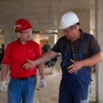 Two construction workers in safety gear discussing plans at a construction site.